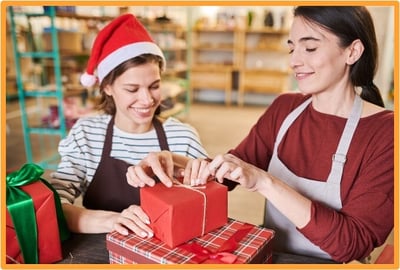 Two women in festive clothing wrapping holiday gifts together, symbolizing charitable giving during the holiday season.
