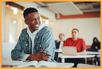 A smiling college student sits at a desk with an open book and pen in a classroom, representing the benefits of saving for higher education with a 529 plan.