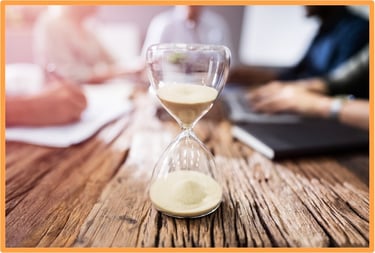Hourglass on a wooden table with people working in the background, symbolizing the approaching deadline for claiming the Work Opportunity Tax Credit.