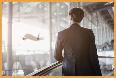 Businessperson standing in an airport terminal looking at a departing airplane, representing business travel and related expense management