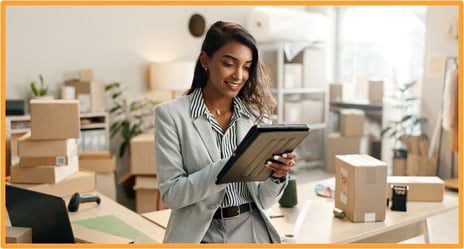 Female entrepreneur using a digital tablet in her small business workspace to organize inventory and manage business growth plans.