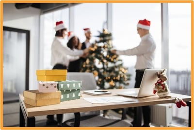 Office decorated for the holidays with wrapped gifts on a desk and colleagues decorating a Christmas tree, representing thoughtful workplace gift-giving.