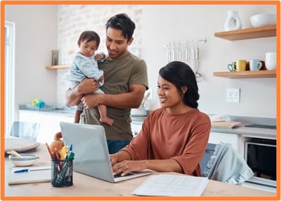 A young family in their kitchen, with one parent holding a baby and the other parent using a laptop, surrounded by paperwork and office supplies.