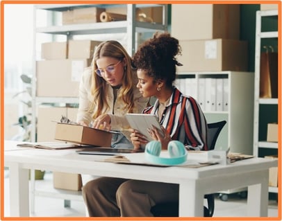Two small business owners preparing a package and reviewing information on a tablet in a storeroom, representing small business operations and tax-saving opportunities for entrepreneurs.