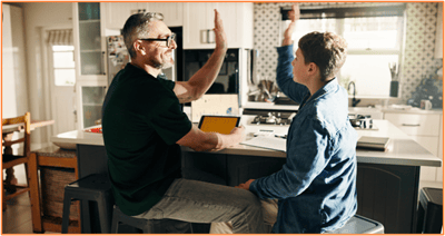 A father and son share a high-five at the kitchen counter while working together with a tablet, highlighting the importance of family collaboration and proactive planning for a child’s future expenses and education.