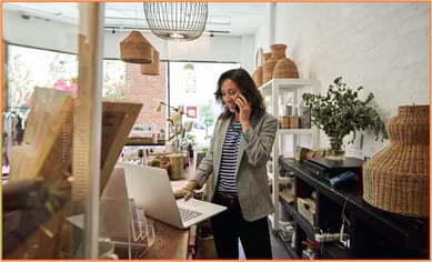 Small business owner talking on the phone while managing finances and retirement contributions on her laptop in her shop.