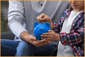 Parent helping a young child put a coin into a blue piggy bank, representing starting early with tax-advantaged savings accounts and teaching children about financial planning.