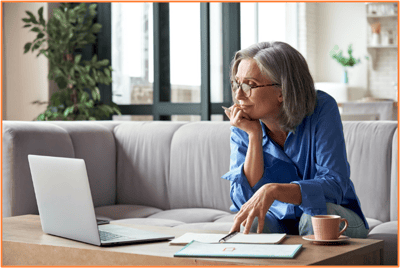 Older woman sitting on a sofa at home, looking thoughtfully at her laptop while taking notes, representing personal retirement planning and consideration of 401(k) options.