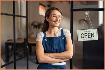 Young entrepreneur standing confidently at the entrance of her new business, smiling next to an open sign, symbolizing the launch of a start-up and the importance of making the right business entity choice.