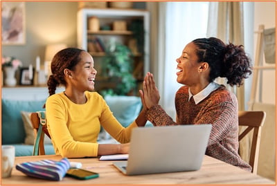 A mother and daughter smile and high-five each other at a table with a laptop and school supplies, representing the teamwork and planning involved in managing the financial challenges of raising a child.