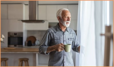 A senior man standing in his kitchen, holding a mug and looking thoughtfully out the window, representing reflection and planning for the future after the loss of a spouse.