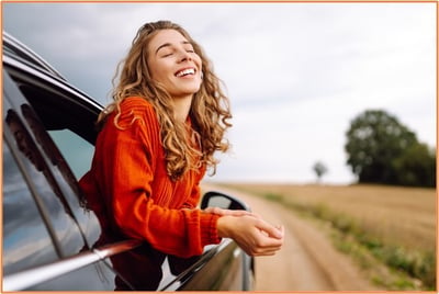 Young woman enjoying a summer road trip, leaning out of a car window on a scenic rural road, symbolizing the joy and freedom of travel.