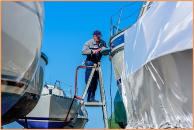 A man standing on a ladder polishing or working on the side of a boat in a marina, with other boats nearby under a clear blue sky.