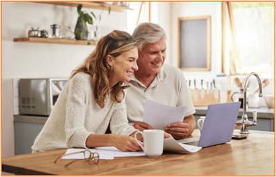 Smiling older couple sitting at a kitchen table, looking at paperwork and a laptop together while planning their finances.