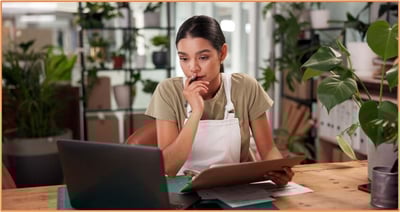 A person in a casual work environment sits at a desk with paperwork and a laptop, appearing thoughtful and focused. This image represents the process of reviewing tax documents and considering deductible interest expenses, such as mortgage, student loan, investment, and auto loan interest, for an individual tax return.