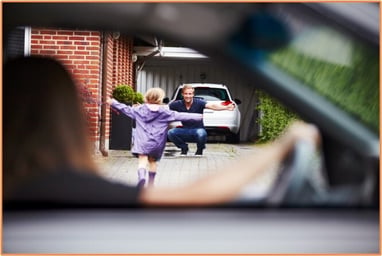 A child runs toward a smiling man waiting by a car in a driveway, viewed from inside another car, illustrating shared parenting and the family transitions that often accompany divorce.