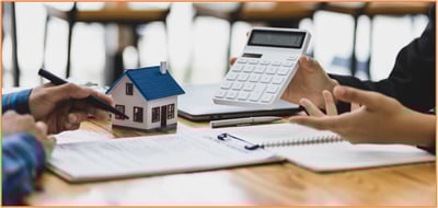 Two individuals review financial documents at a desk, with a model house and a large calculator in the foreground. This scene symbolizes calculating mortgage interest deductions and discussing the requirements for interest expense write-offs related to home loans on a federal income tax return.
