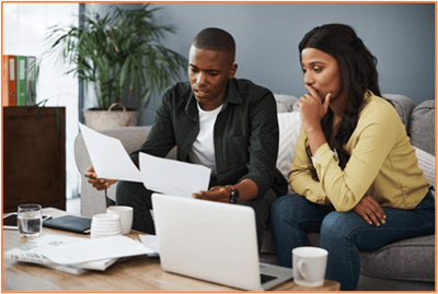 A couple sits on a couch looking over financial documents and a laptop, appearing serious and thoughtful, representing the financial challenges and important decisions involved in divorce planning.