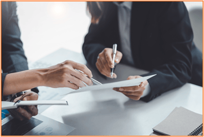 Close-up of two people in business attire discussing and reviewing a document together at a desk, with pens and paperwork visible.