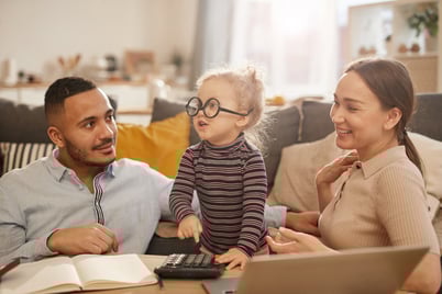 A young family sits together in a cozy living room, smiling and engaging with their small child, who is playfully wearing oversized glasses and using a calculator. The scene suggests family financial planning and highlights the importance of tax breaks and savings options for families, as discussed in the article about the new "One, Big, Beautiful Bill Act" and its expanded tax benefits for families with children and dependents.