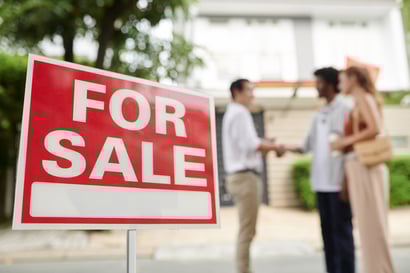A red "For Sale" sign is displayed in front of a house, with three people in the background shaking hands, representing a home selling or buying transaction.