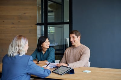 A couple sits at a table having a positive discussion with a professional advisor, reviewing documents and a tablet. This scene represents homeowners seeking expert guidance on their options and the tax implications when selling a home in a challenging real estate market.