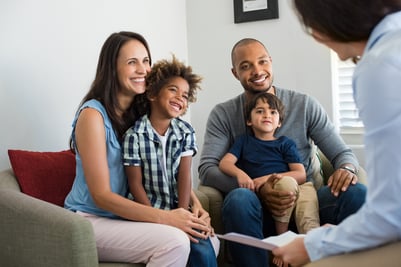 A smiling family with two young children sits together on a couch, attentively listening to a professional. The scene represents families seeking guidance about new tax breaks and financial planning opportunities under the One, Big, Beautiful Bill Act (OBBBA) discussed in the blog.